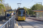 A yellow tram approaches a station with passengers waiting on the platform under clear blue skies.